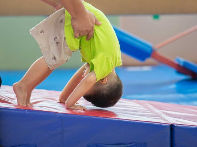 Child in a green shirt and shorts doing a headstand on a blue and red mat, assisted by an adult.
