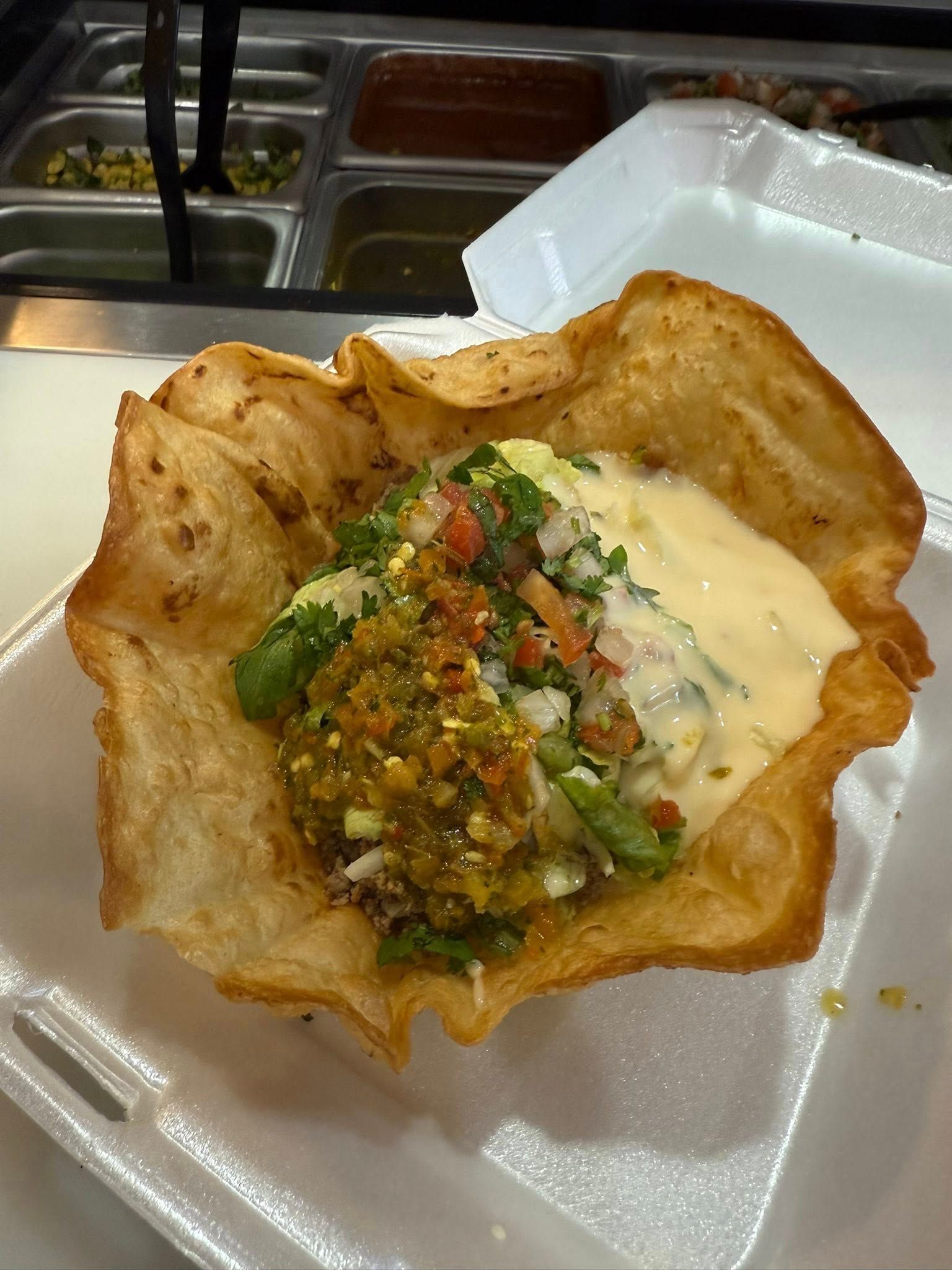 Taco salad in a crispy shell, filled with toppings and cheese sauce, in a styrofoam container.