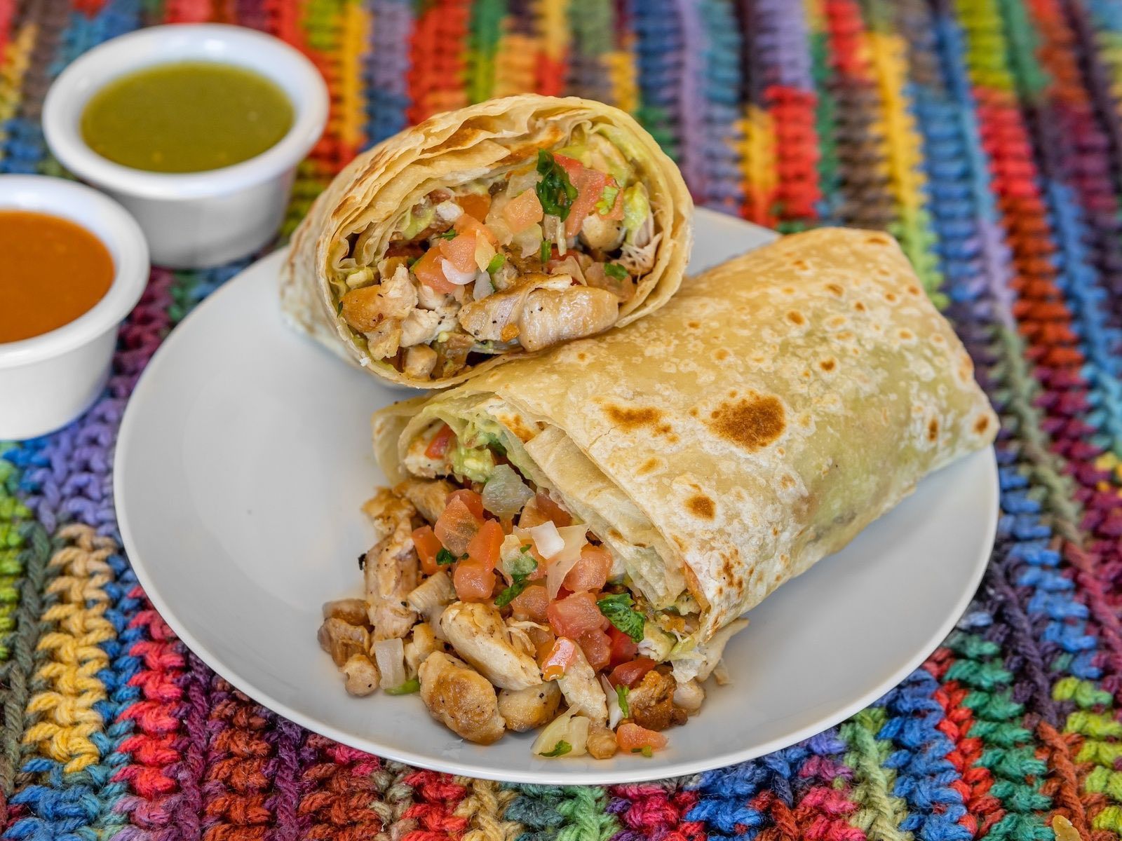 Burrito cut open on a white plate, with salsa in small cups, on a colorful woven mat.