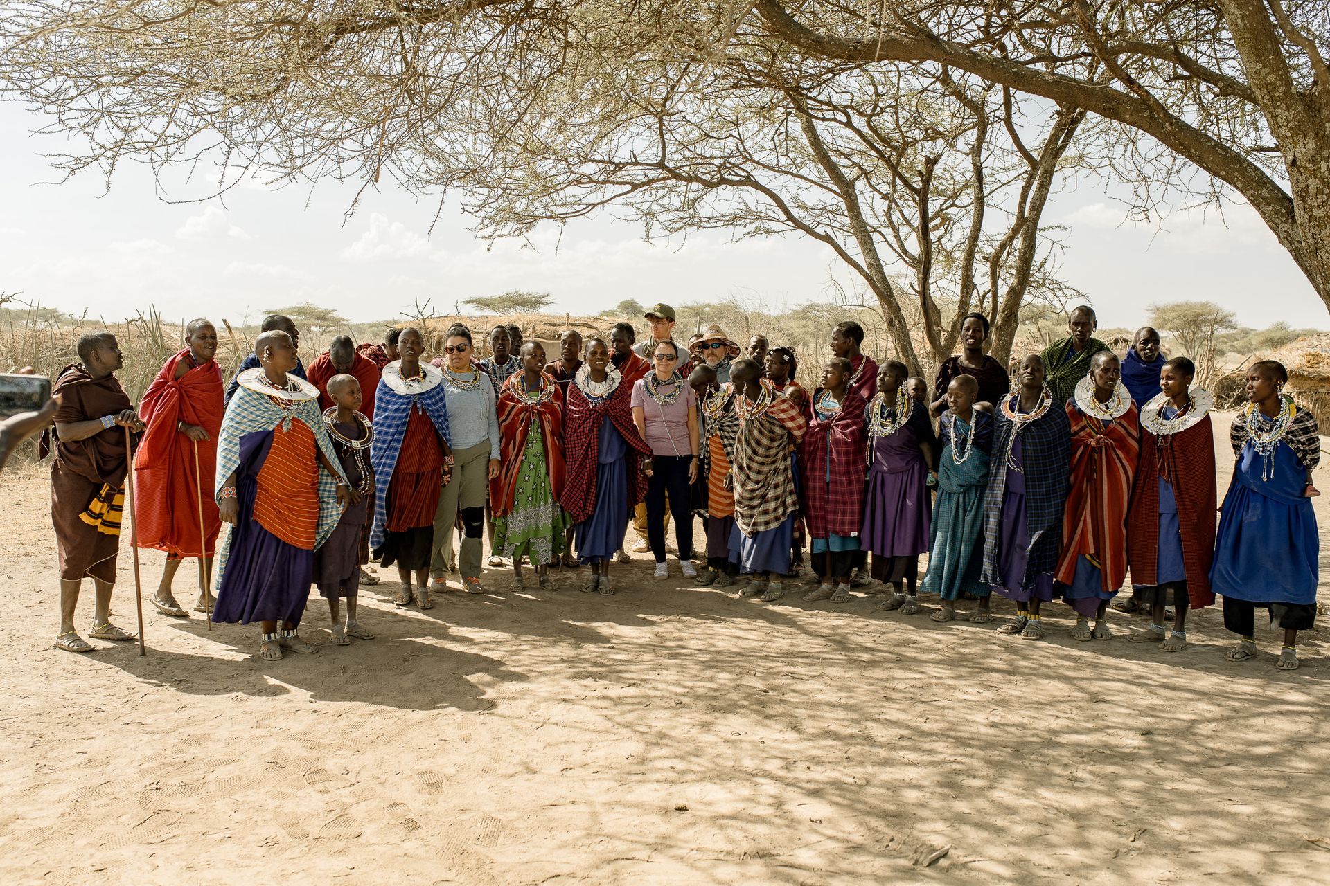 A large group of people are standing under a tree in the desert.