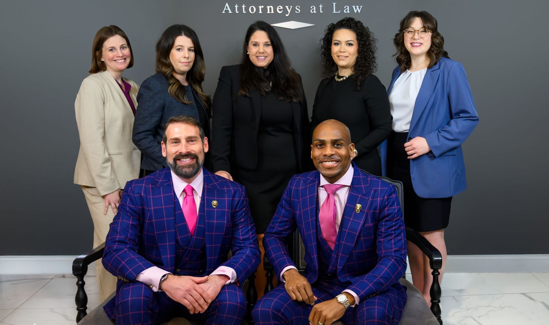 Law firm staff poses for a photo: two men seated, six women standing, dark suits, professional setting.