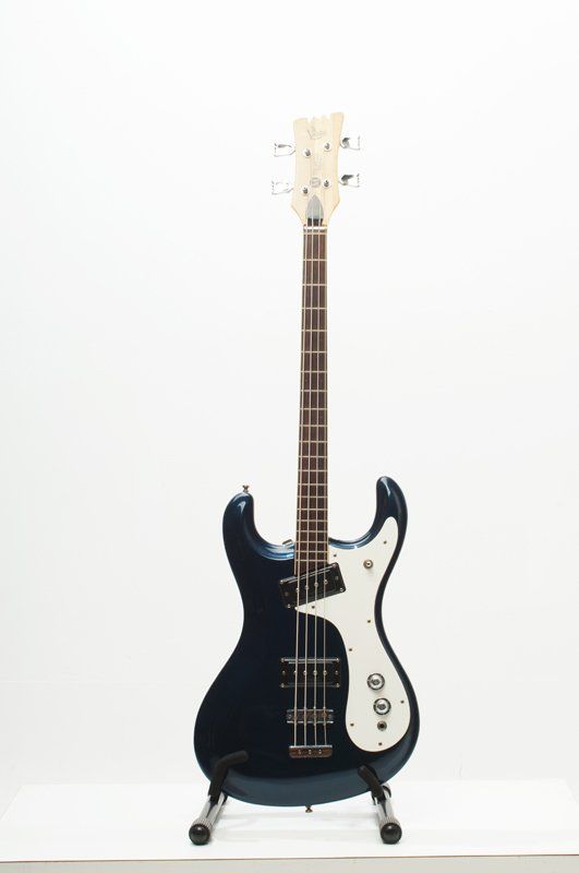 A blue electric bass guitar is sitting on a stand on a white background.