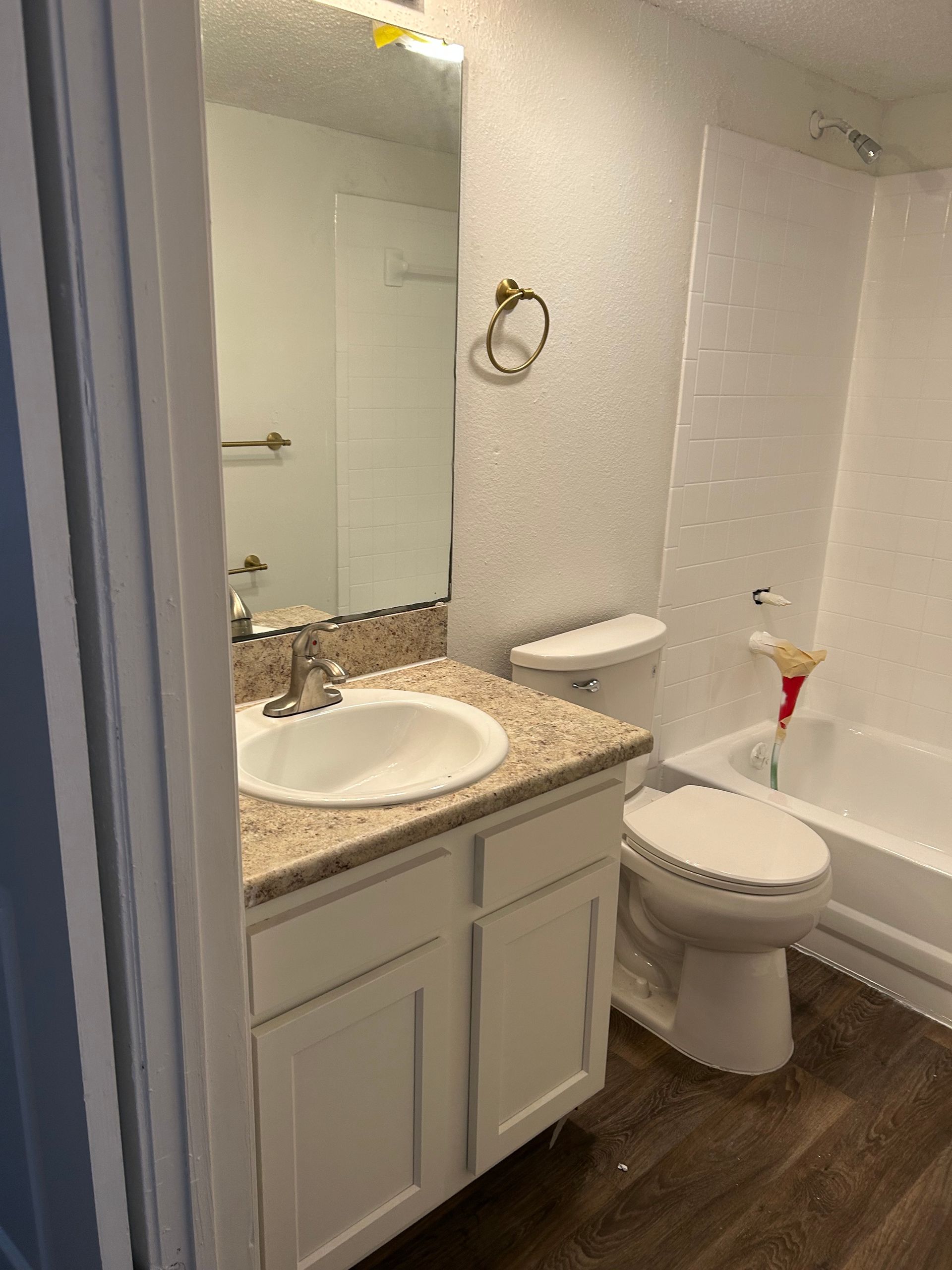 Bathroom with white vanity, toilet, and bathtub. Beige countertop, gold towel ring, and brown flooring.
