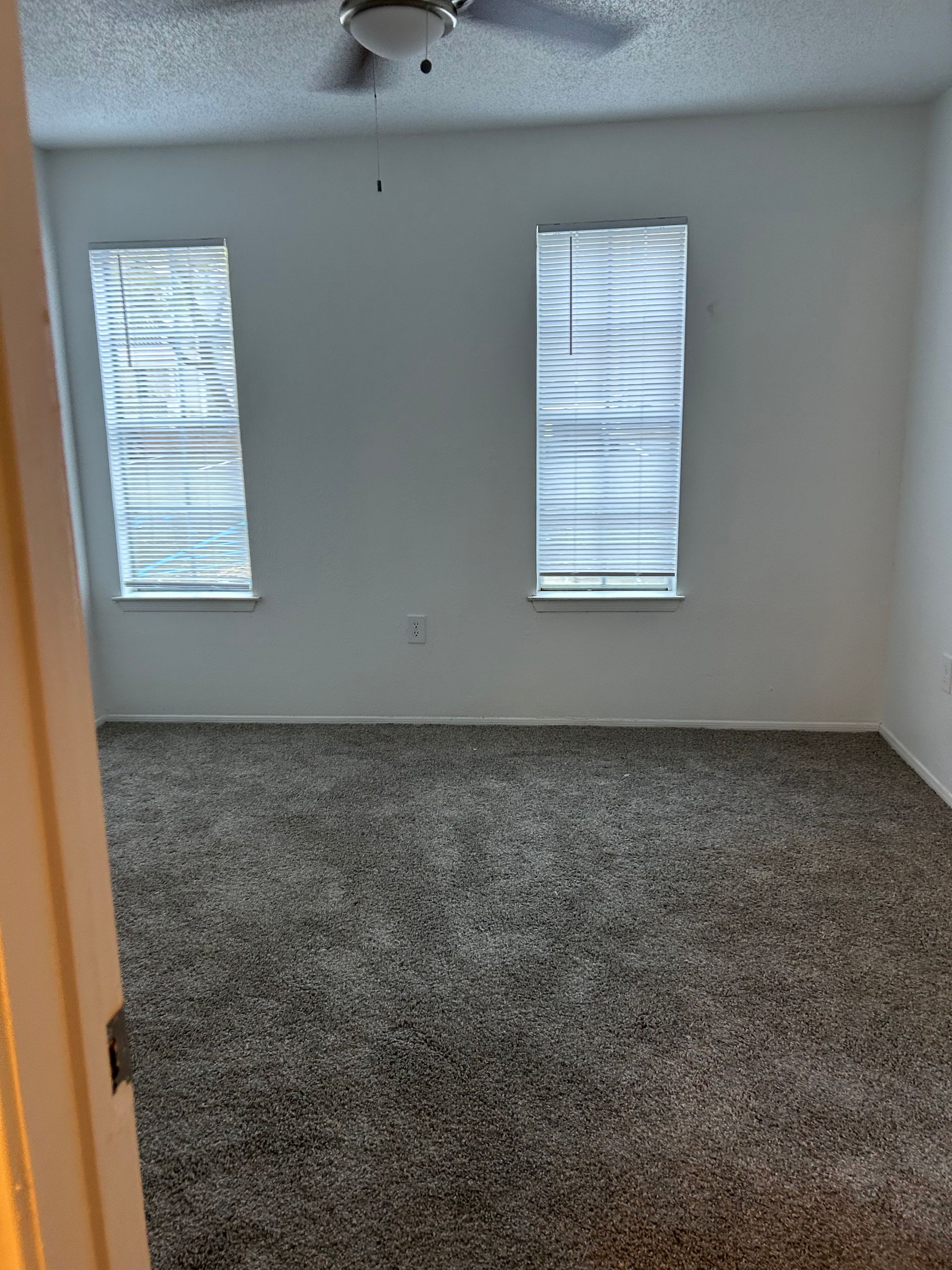 Empty room with gray carpet, two windows with blinds, white walls, and a ceiling fan.