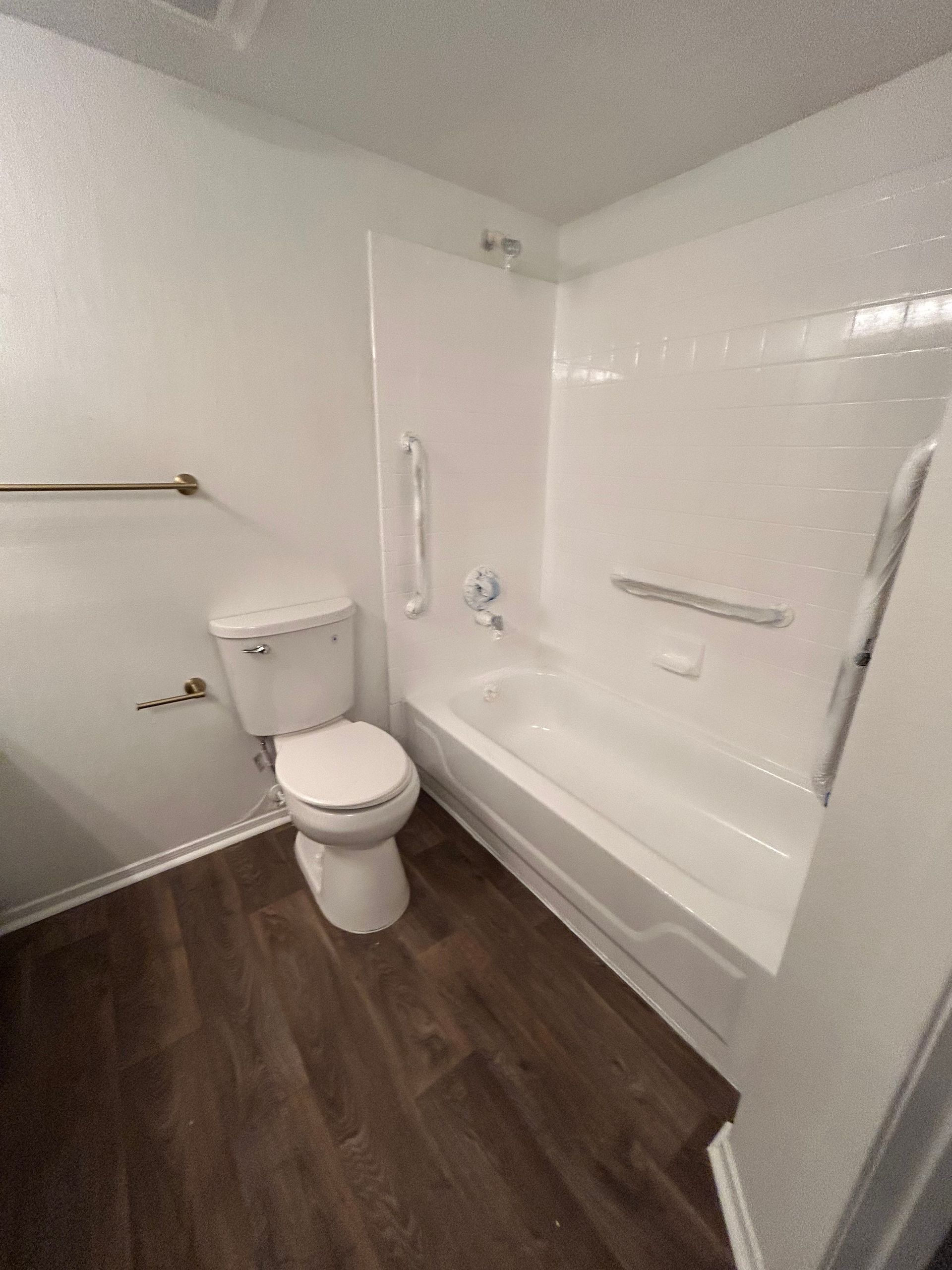 Bathroom with white fixtures, a toilet, tub with grab bars, and dark wood-look flooring.