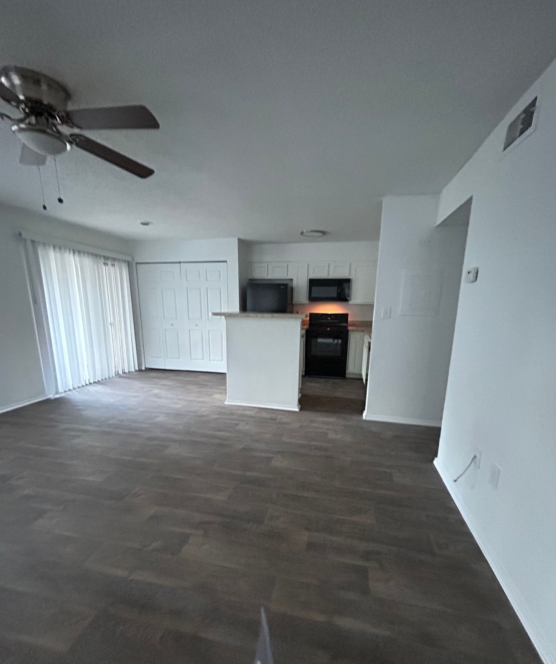 Empty living space with wood-look flooring, white walls, and a kitchen visible in the back.