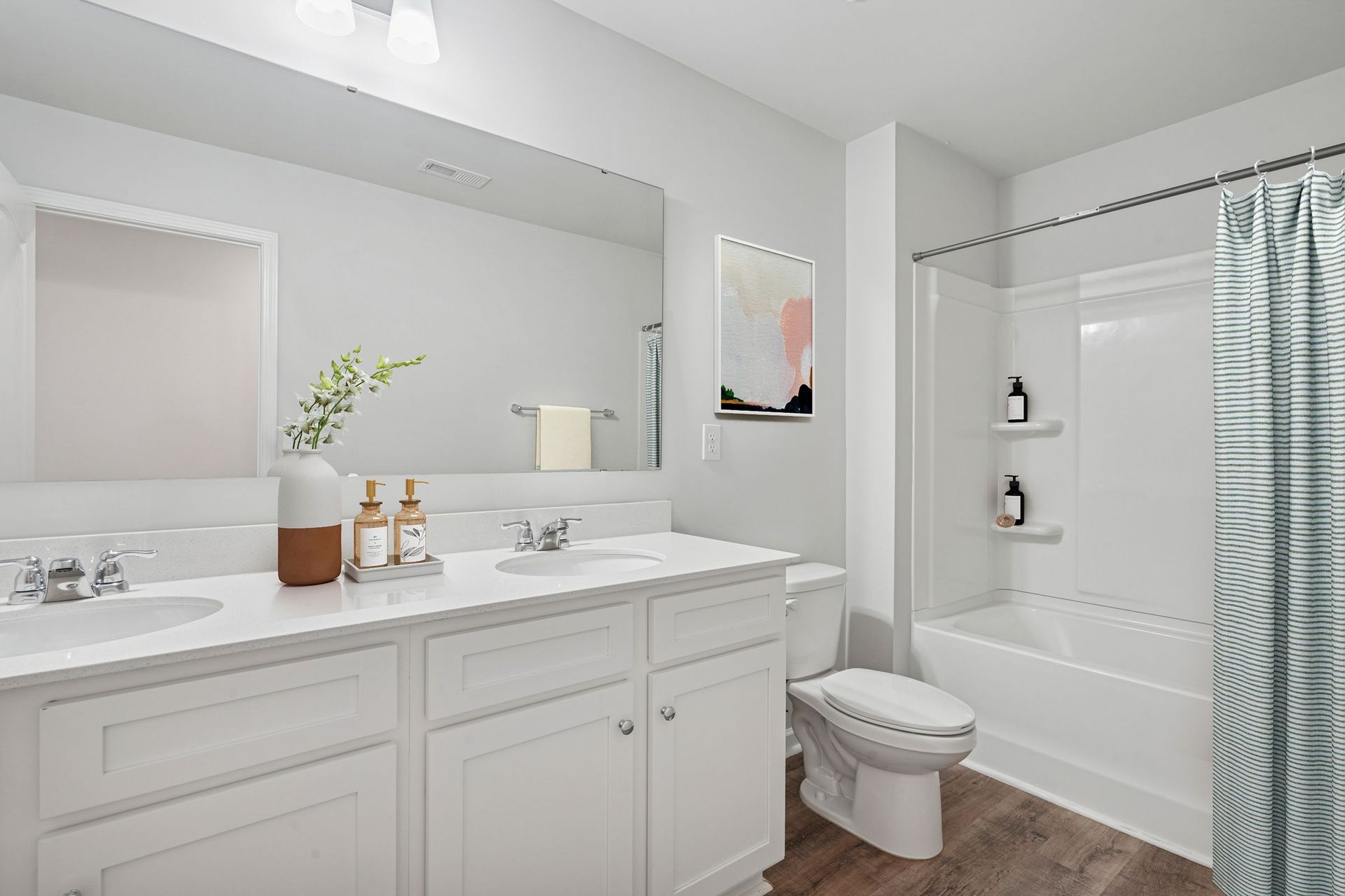 White bathroom with double vanity, bathtub with shower curtain, and a framed art piece.