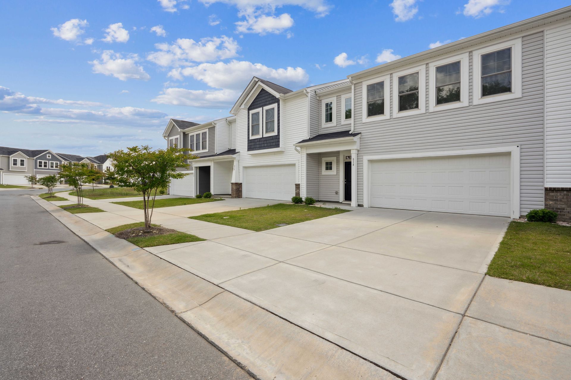 Row of modern townhouses with white siding, driveways, and green lawns on a street under a cloudy sky.