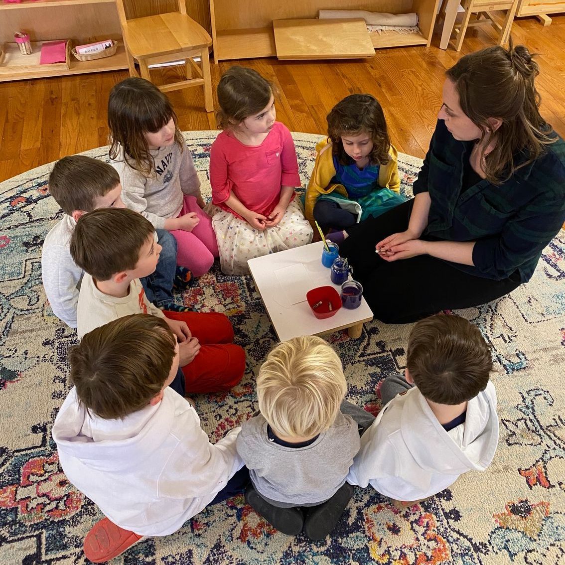 A group of children are sitting in a circle on the floor with a teacher in a montessori classroom.