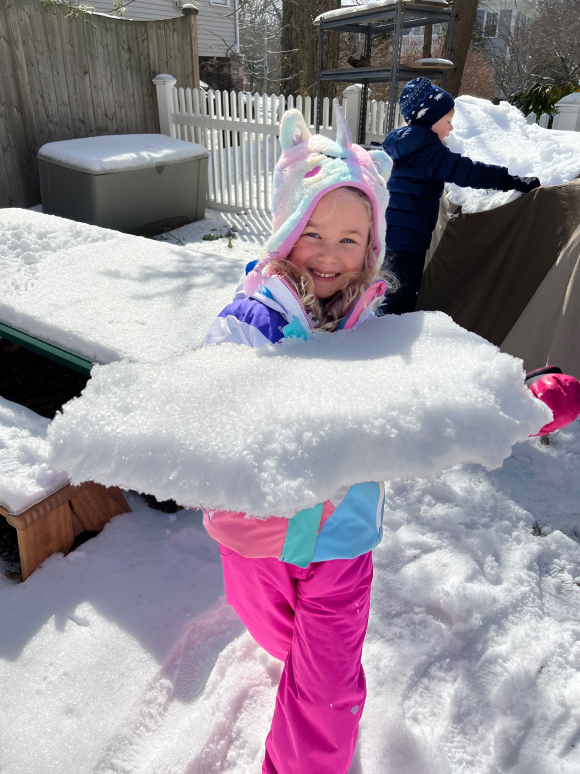 Montessori child smiling, holding large snow slab in snowy backyard.