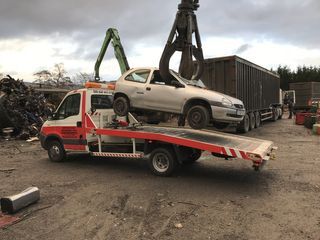 Une dépanneuse soulevant une voiture argentée à l'aide d'une pince métallique ; décor d'une casse automobile.