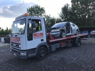 Dépanneuse avec une voiture argentée endommagée sur son plateau. Extérieur, ciel nuageux.