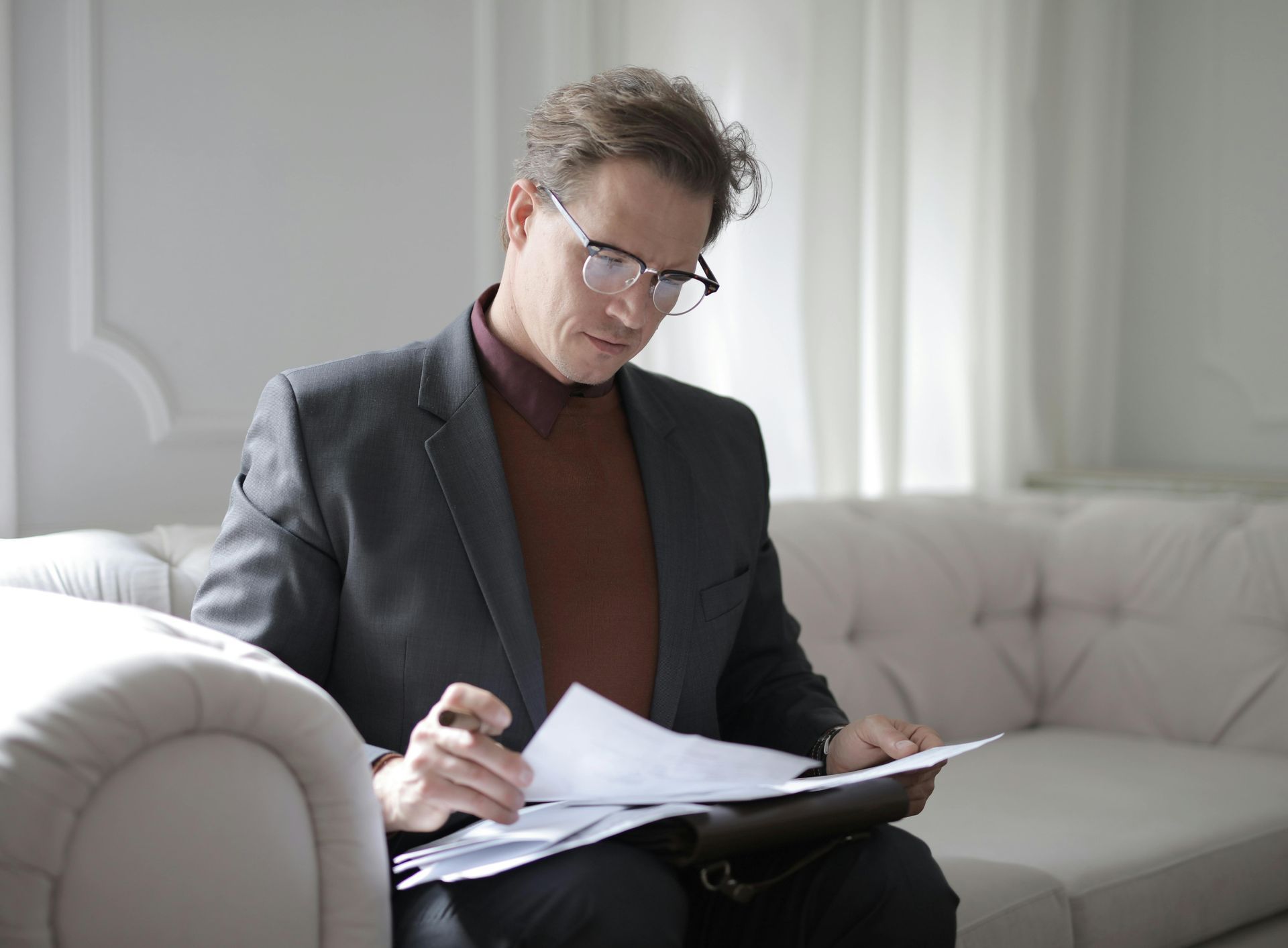 Un homme portant des lunettes et un costume examine des documents, assis sur un canapé couleur crème.