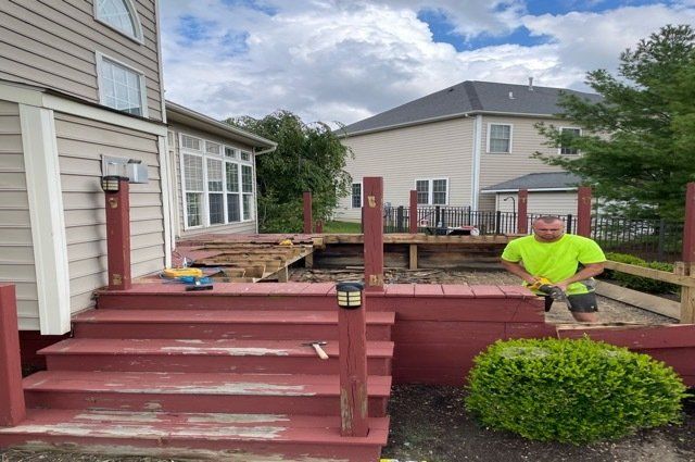 Red deck in backyard that is halfway torn down. Guy on the right with a saw cutting up part of the deck.