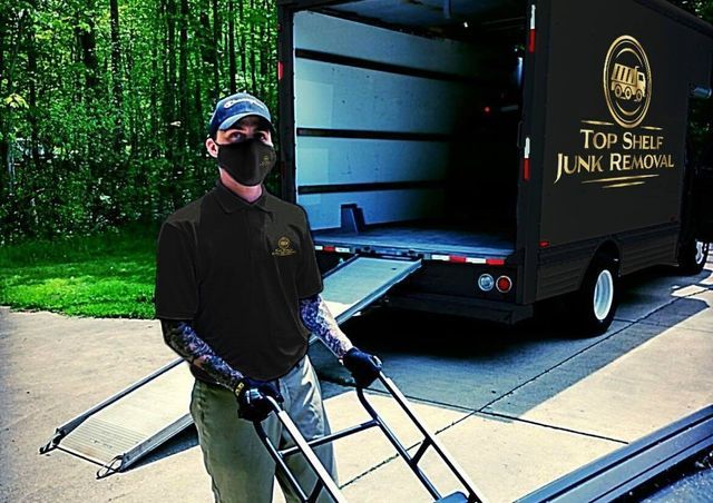 a man with tattoos holding a dolly wearing a top shelf junk removal shirt standing next to a black box truck