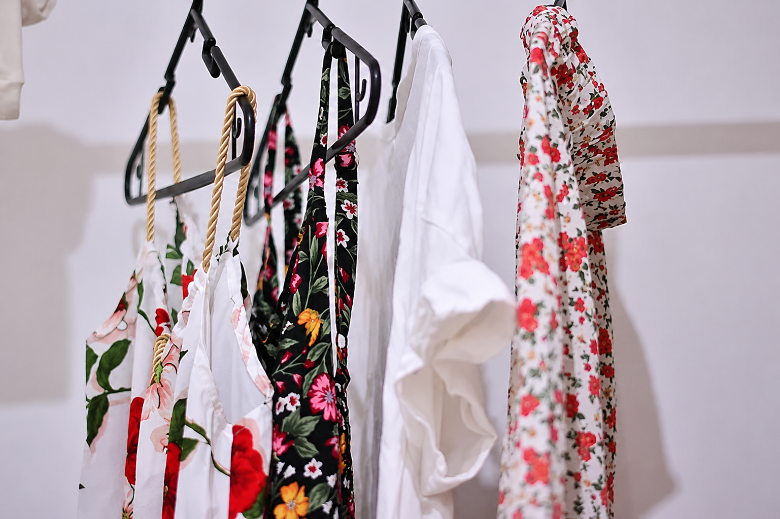 Floral dresses and a white blouse hanging on black hangers against a light wall