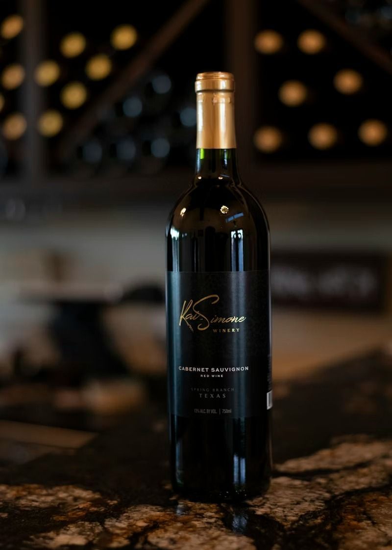A dark wine bottle with a gold label and cap on a marble table, with blurred wine bottles in the background.