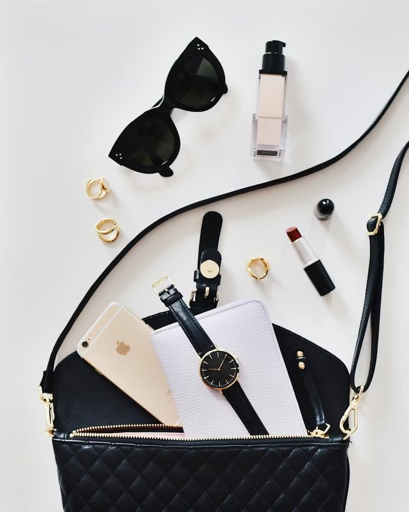 Black quilted handbag with sunglasses, makeup, gold jewelry, watch, notebook, phone, and lipstick on a white background