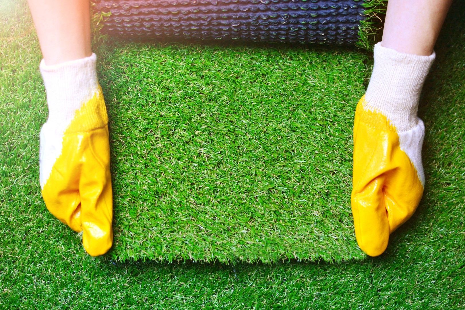 Person cutting artificial grass with a utility knife, measuring with a tape measure on wooden floor.