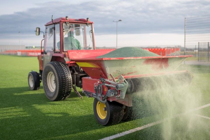 Tractor spreading green pellets on a turf field. Operator wearing protective gear.