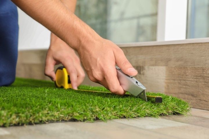 Person cutting artificial grass with a utility knife, measuring with a tape measure on wooden floor.