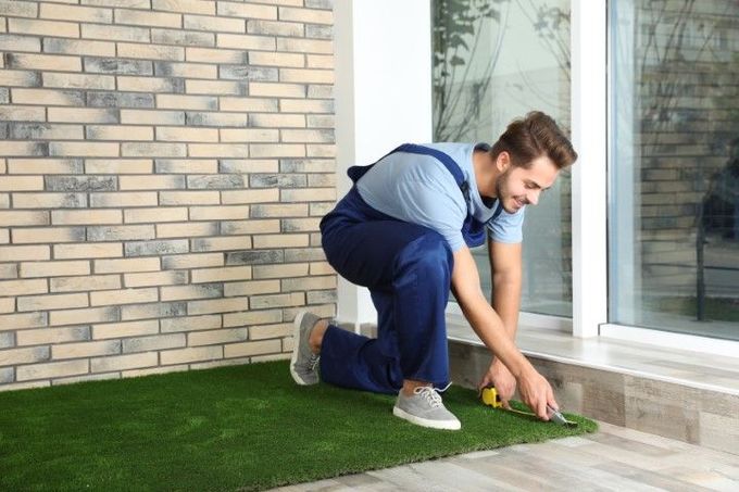 Man in blue overalls cutting artificial turf on a porch with brick wall and glass door.