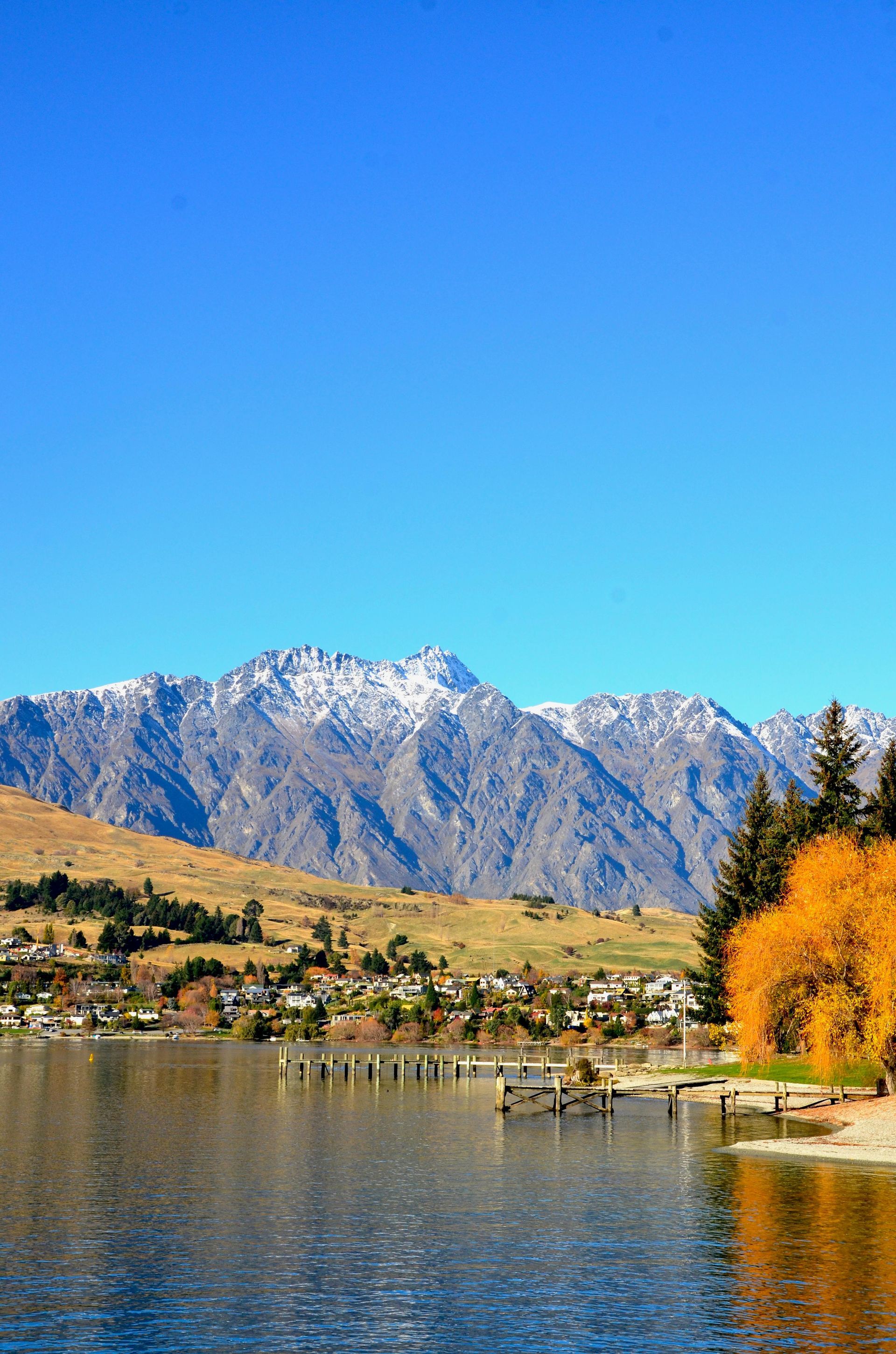 Snow-capped mountains over a calm lake, with a town along the shore and bright blue sky.