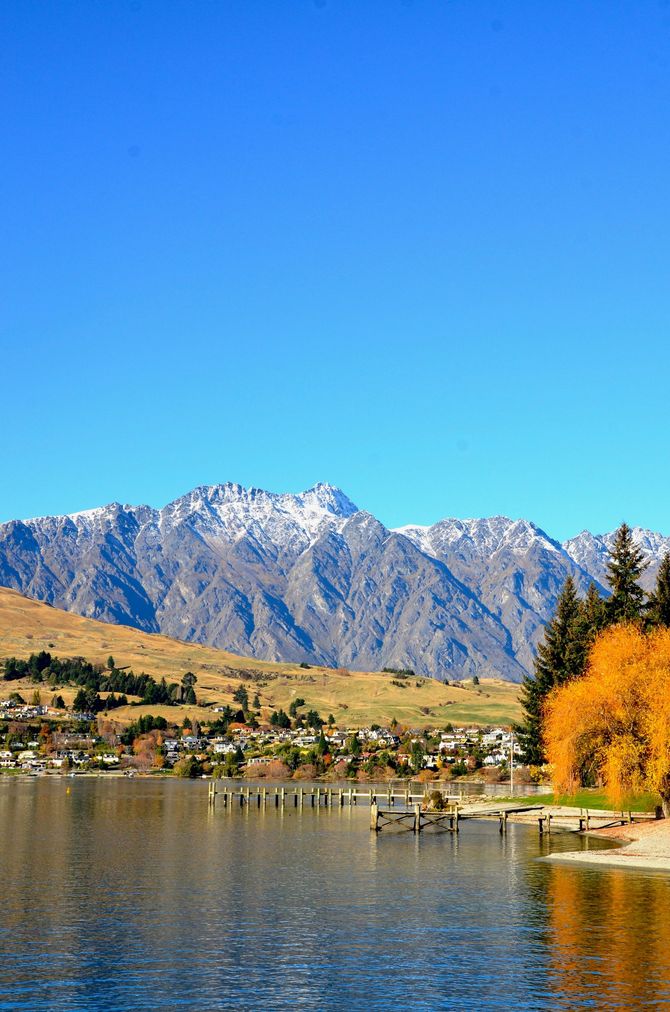 Snow-capped mountains over a calm lake, with a town along the shore and bright blue sky.