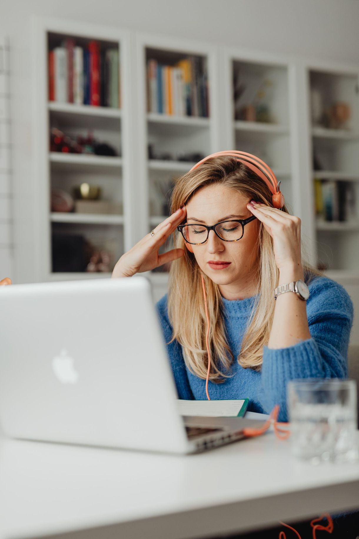 A woman working at a laptop, holding her temples at a desk in a bright office.