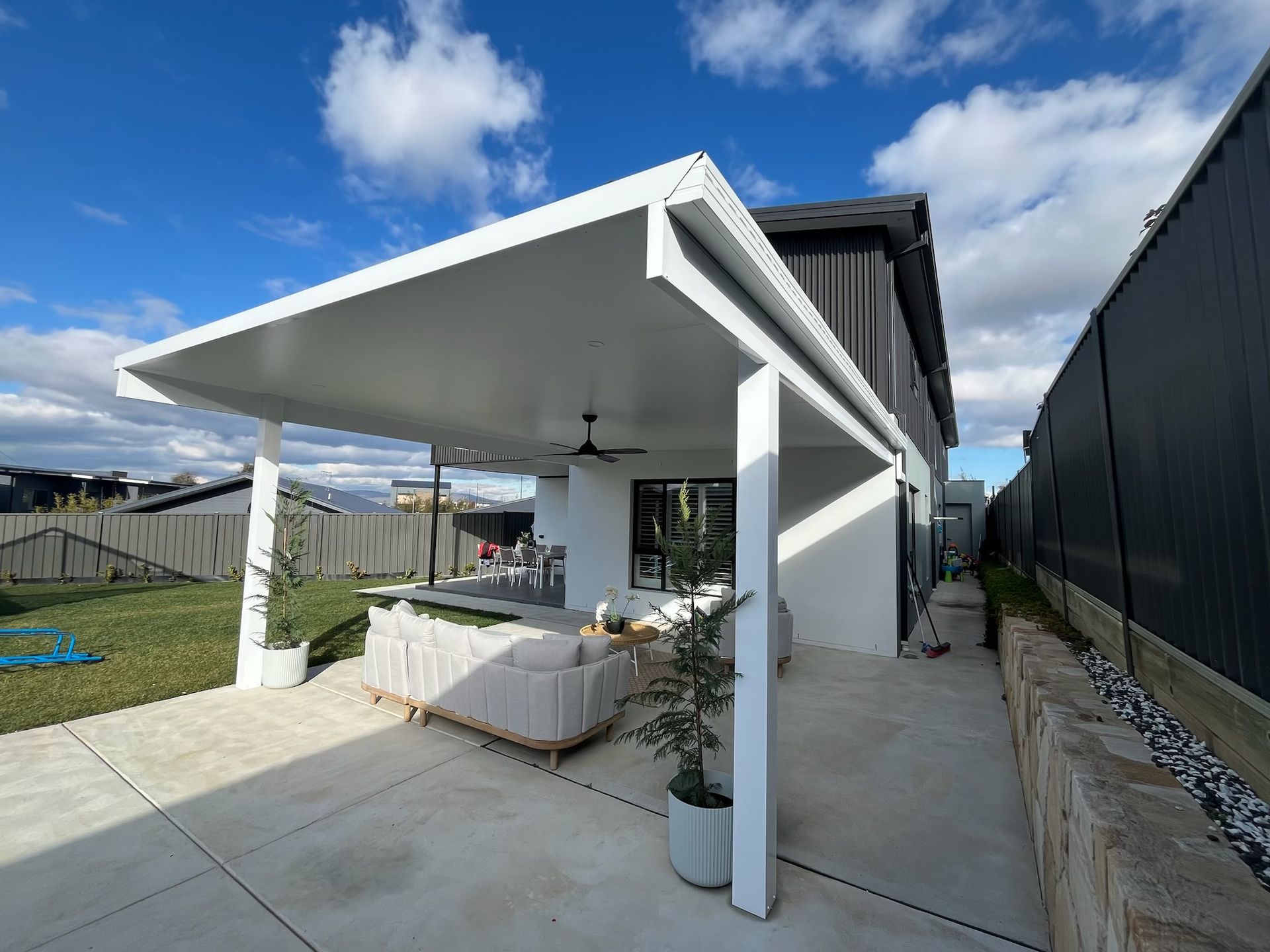 Backyard patio with white roof, concrete, couch, and view of house and fence on sunny day. — Hallam & Sons in Beard, ACT