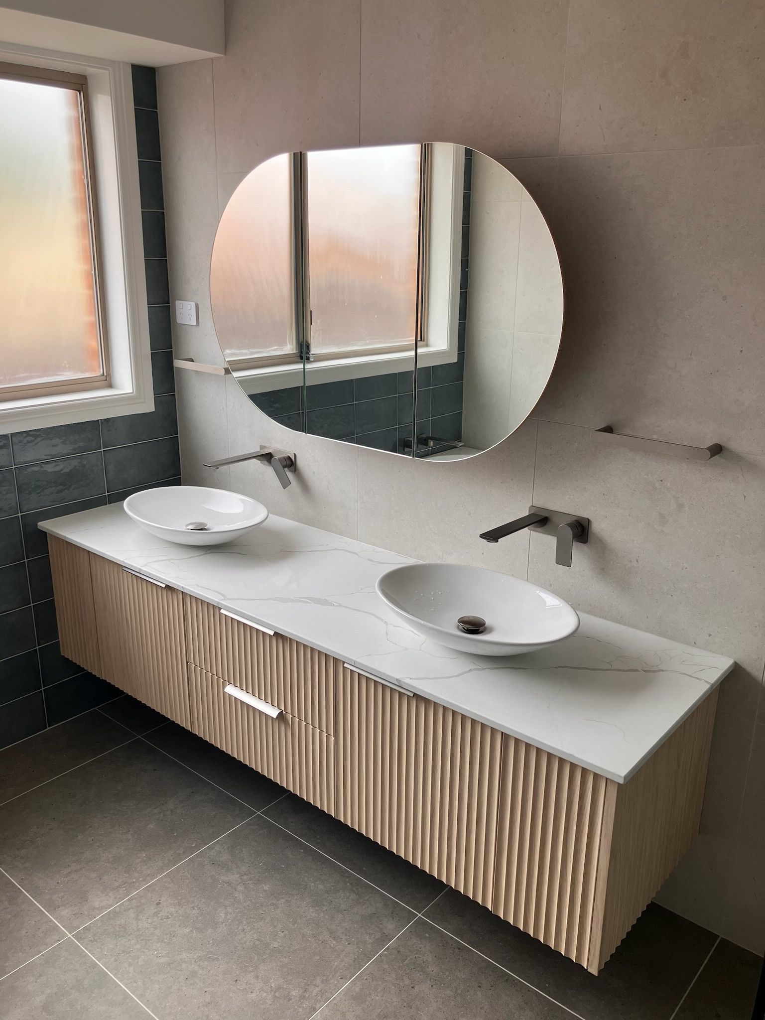 Modern bathroom with a light wood vanity, white countertop, two vessel sinks, and a large oval mirror. — Hallam & Sons in Beard, ACT