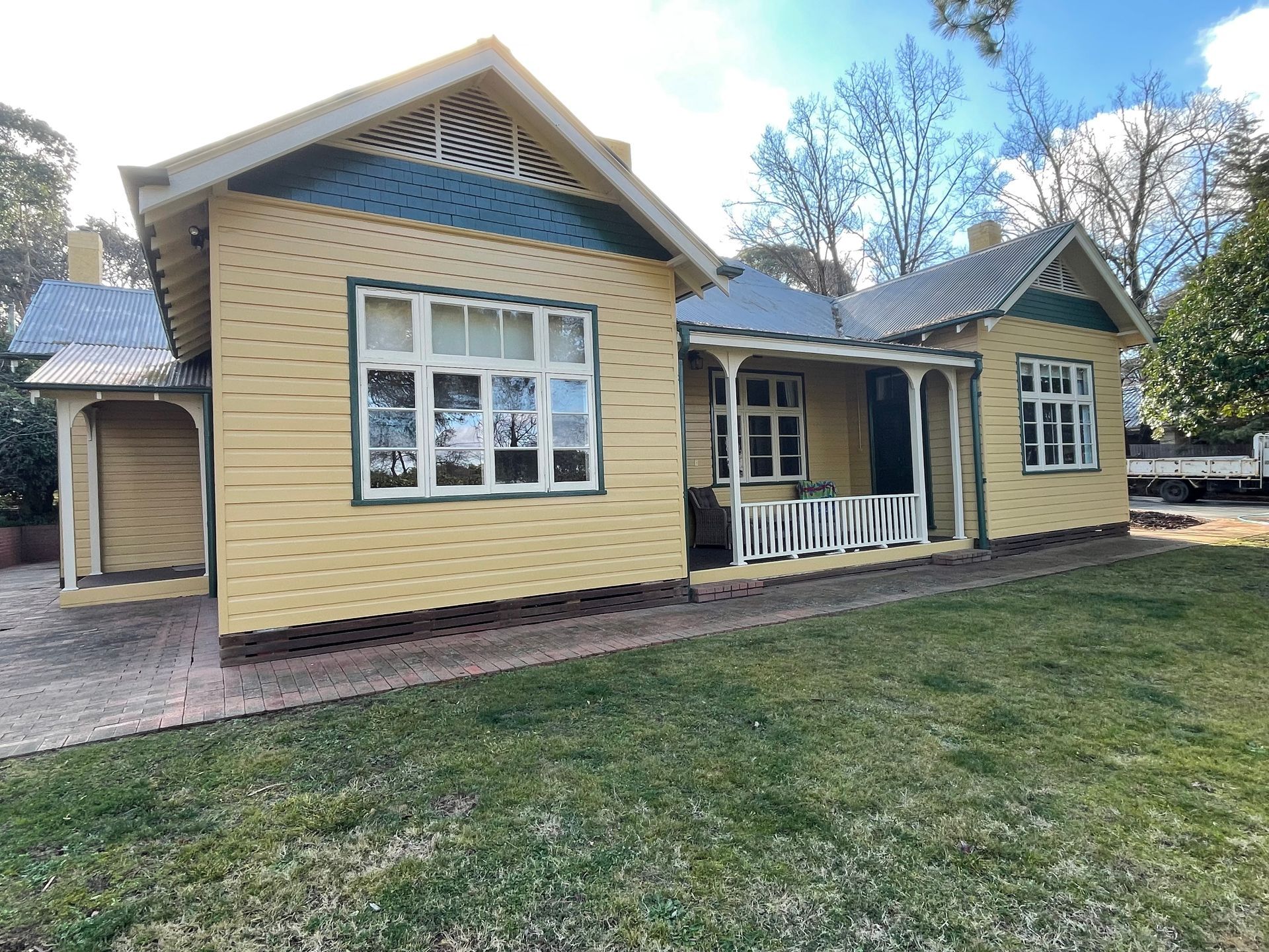 Yellow wooden house with a porch and white-framed windows, set on a green lawn with trees in the background. — Hallam & Sons in Beard, ACT