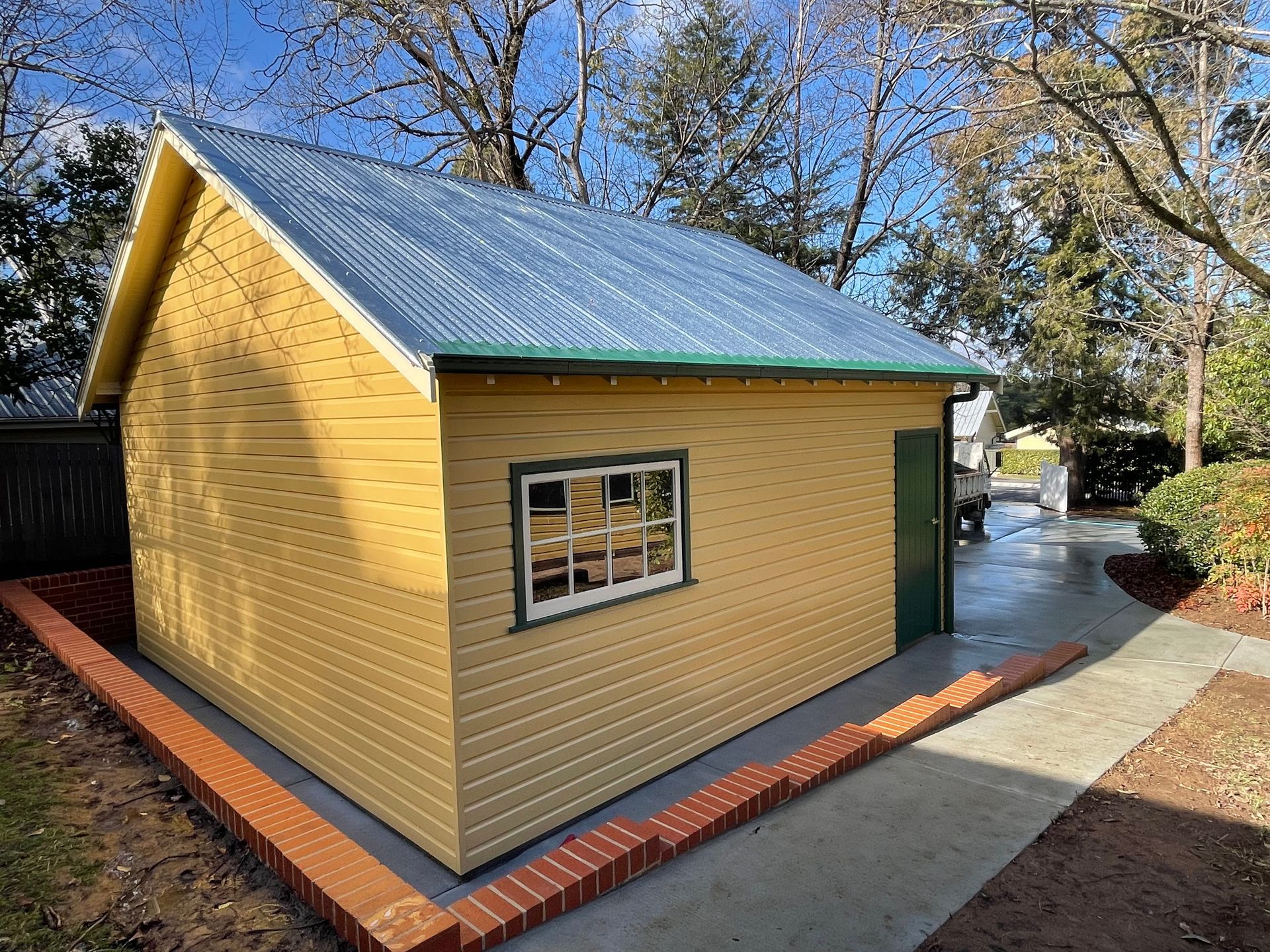 Yellow shed with a corrugated metal roof, window, and green door. Brick border surrounds its base. — Hallam & Sons in Beard, ACT