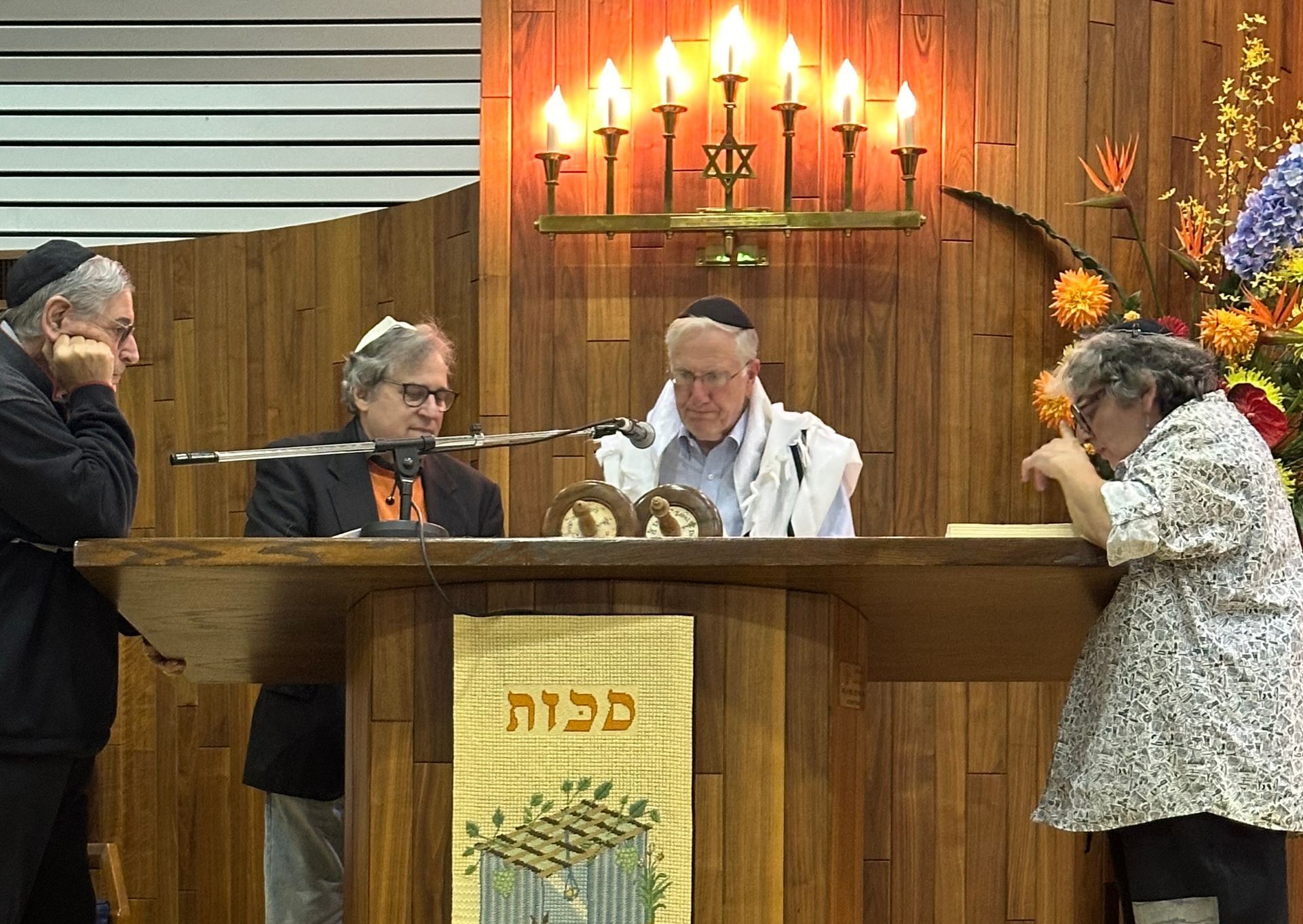 Two men are standing in front of a wall with hebrew writing on it.