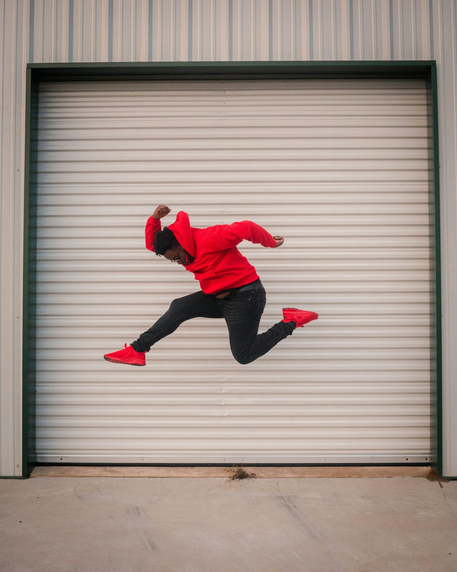 A man in a red hoodie is jumping in the air in front of a garage door.