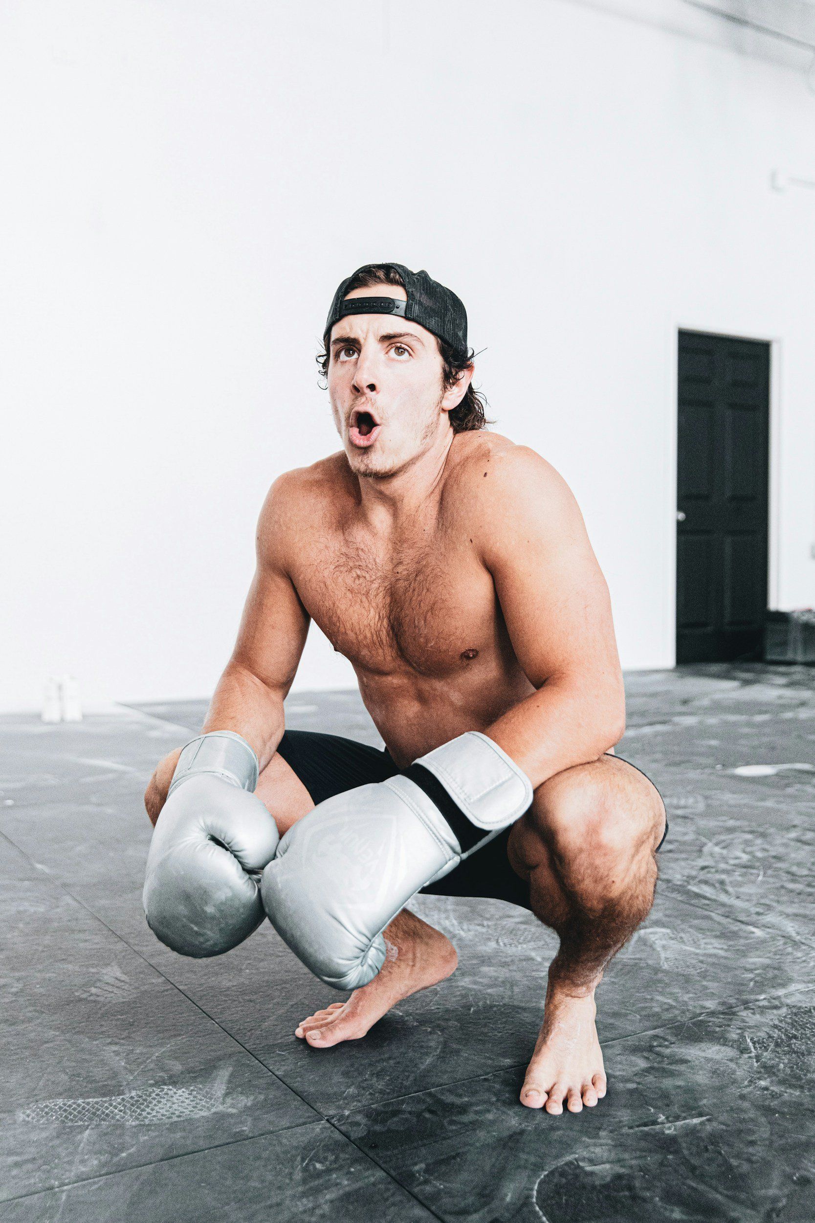 A shirtless man is squatting down with boxing gloves on.