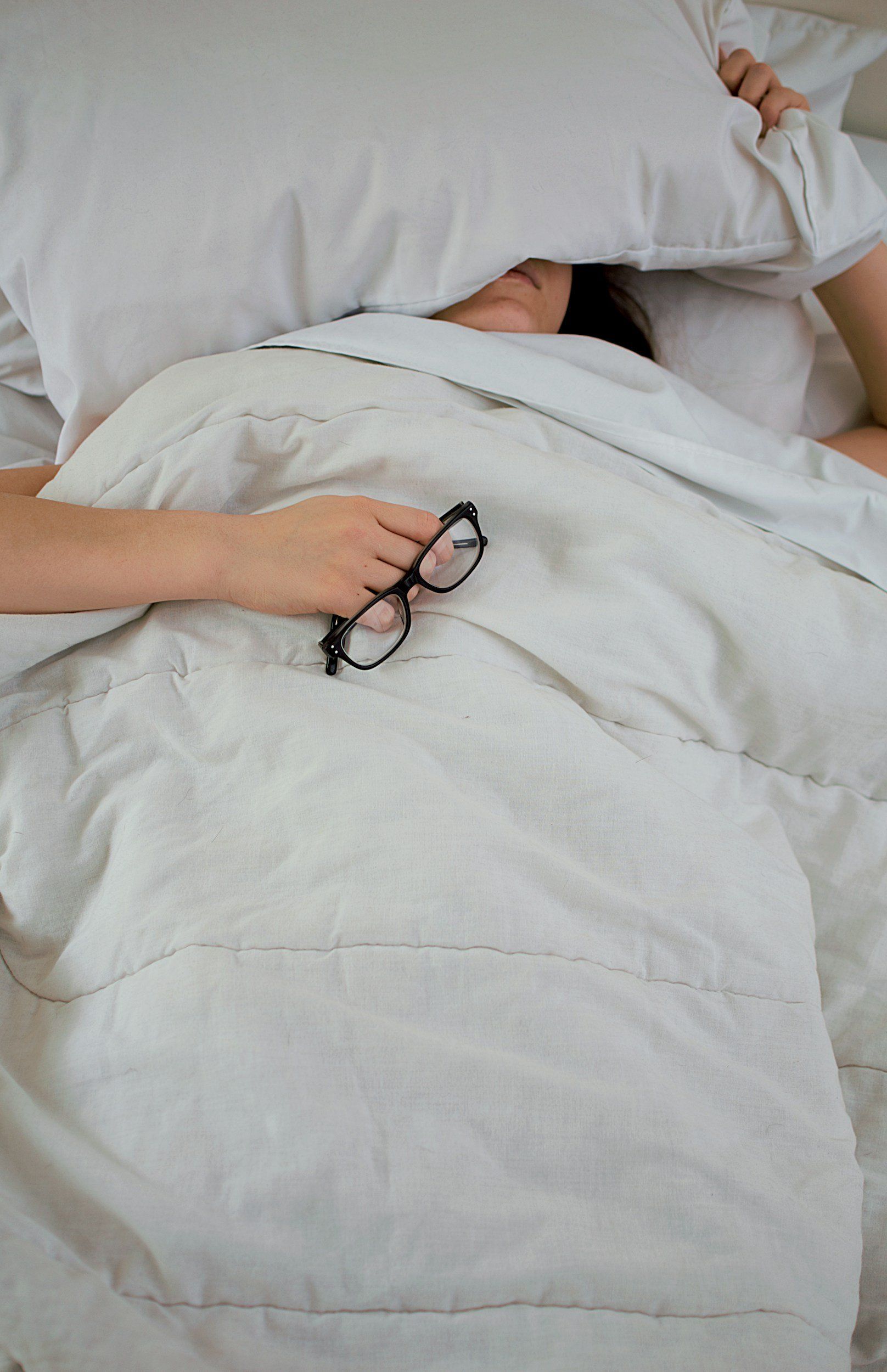 A woman is laying in bed covering her face with a pillow and holding a pair of glasses.