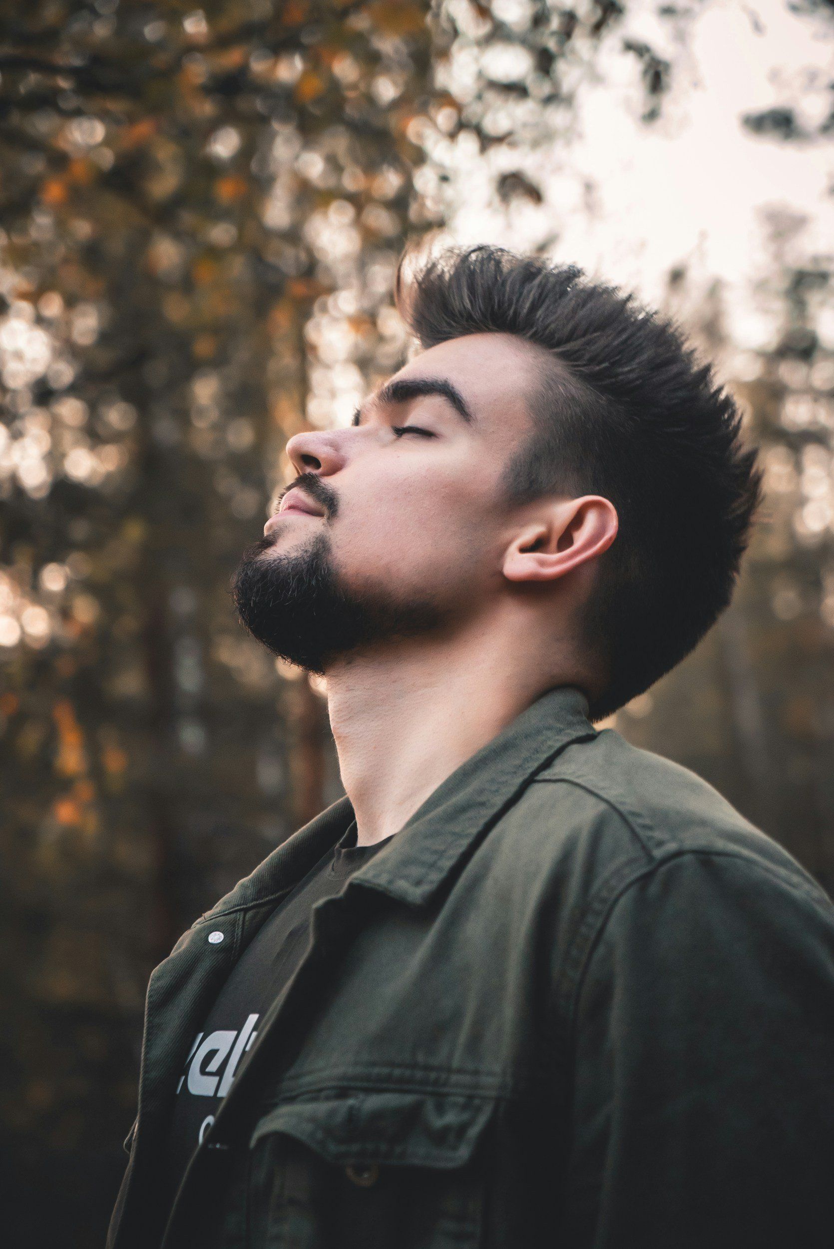 A man with a beard is standing in the woods with his eyes closed.
