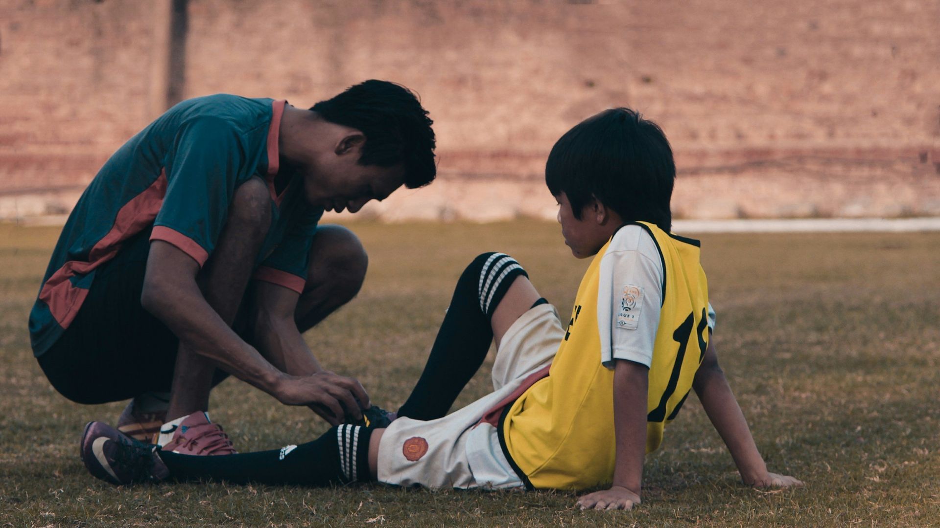A man is helping a young boy with his soccer shoes on a field.