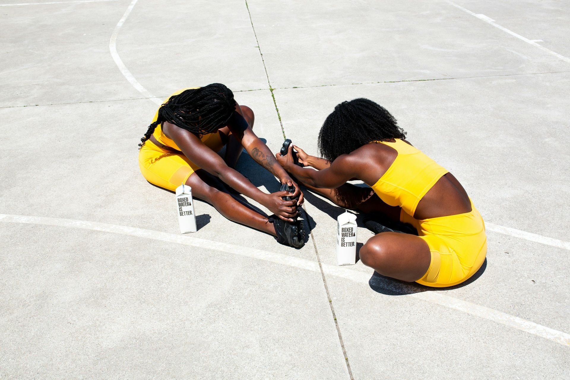 Two women are sitting on the ground stretching their legs on a basketball court.
