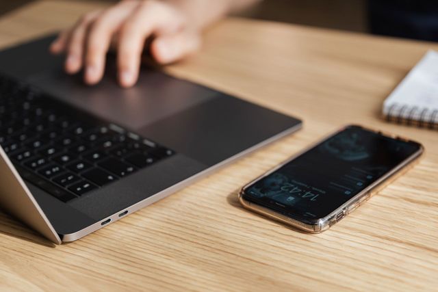 A person is typing on a laptop next to a cell phone.