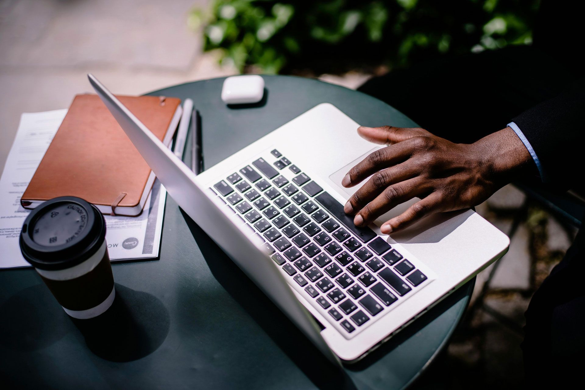 A person's hand on a laptop keyboard. Coffee, notebook, and wireless earbuds on a green table outdoors.