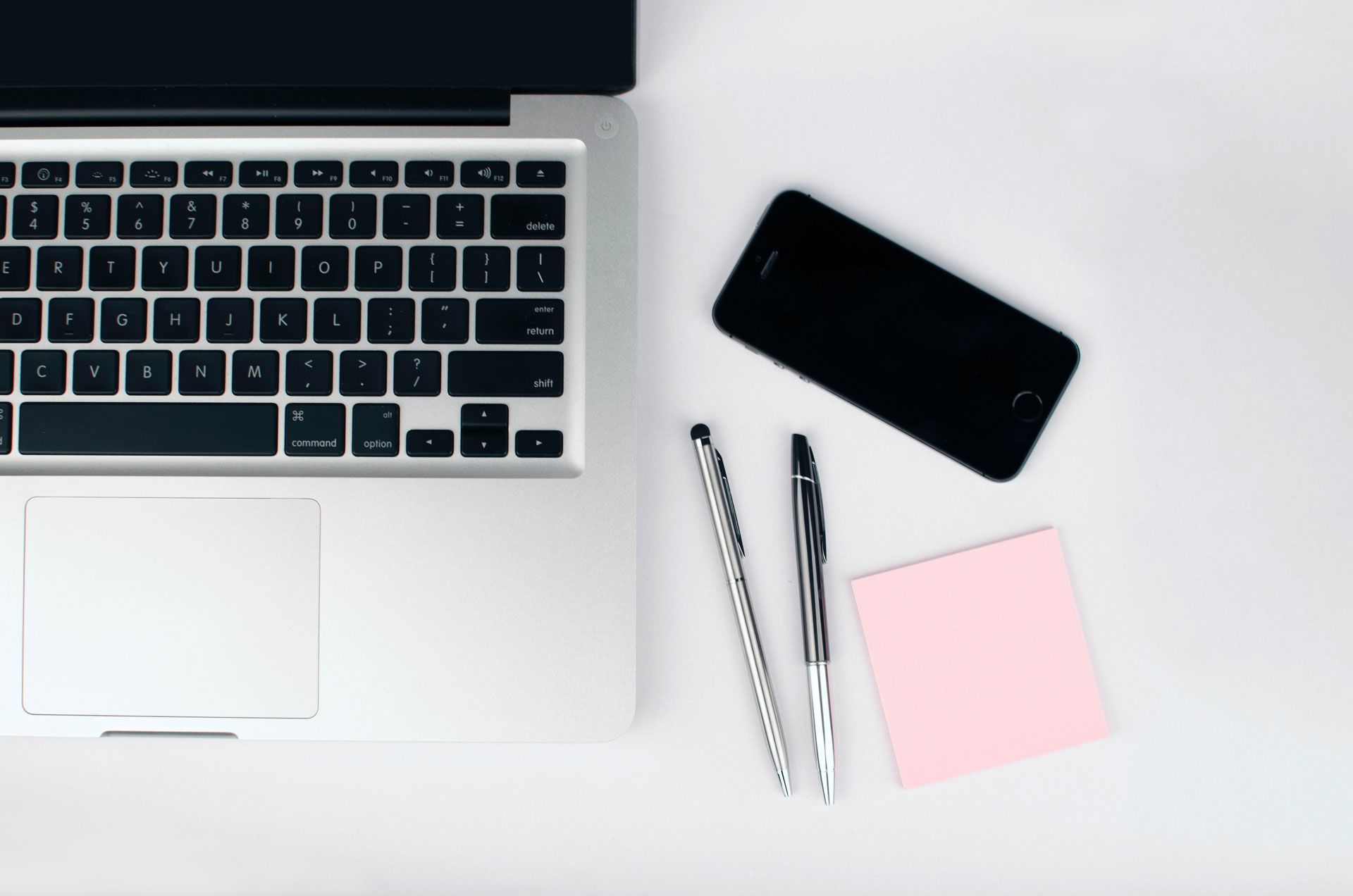 A laptop , cell phone , pen and sticky notes on a white table.