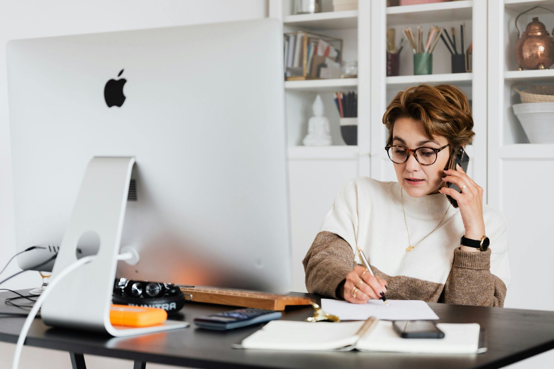 Woman on phone, writing notes at desk with a computer and phone. White shelves in background.