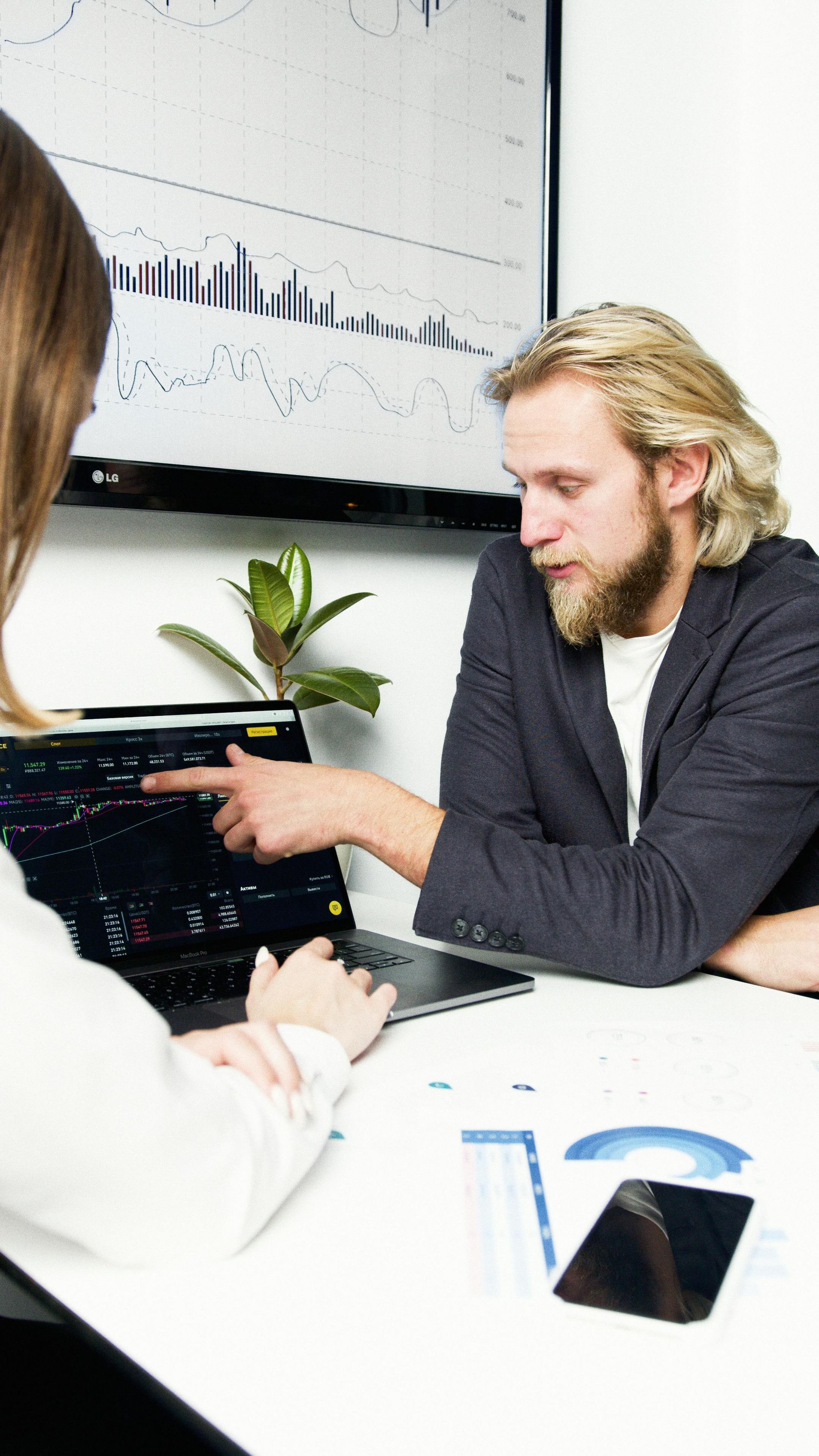 Two people collaborate in an office, reviewing stock market charts on a laptop and a large wall-mounted screen.