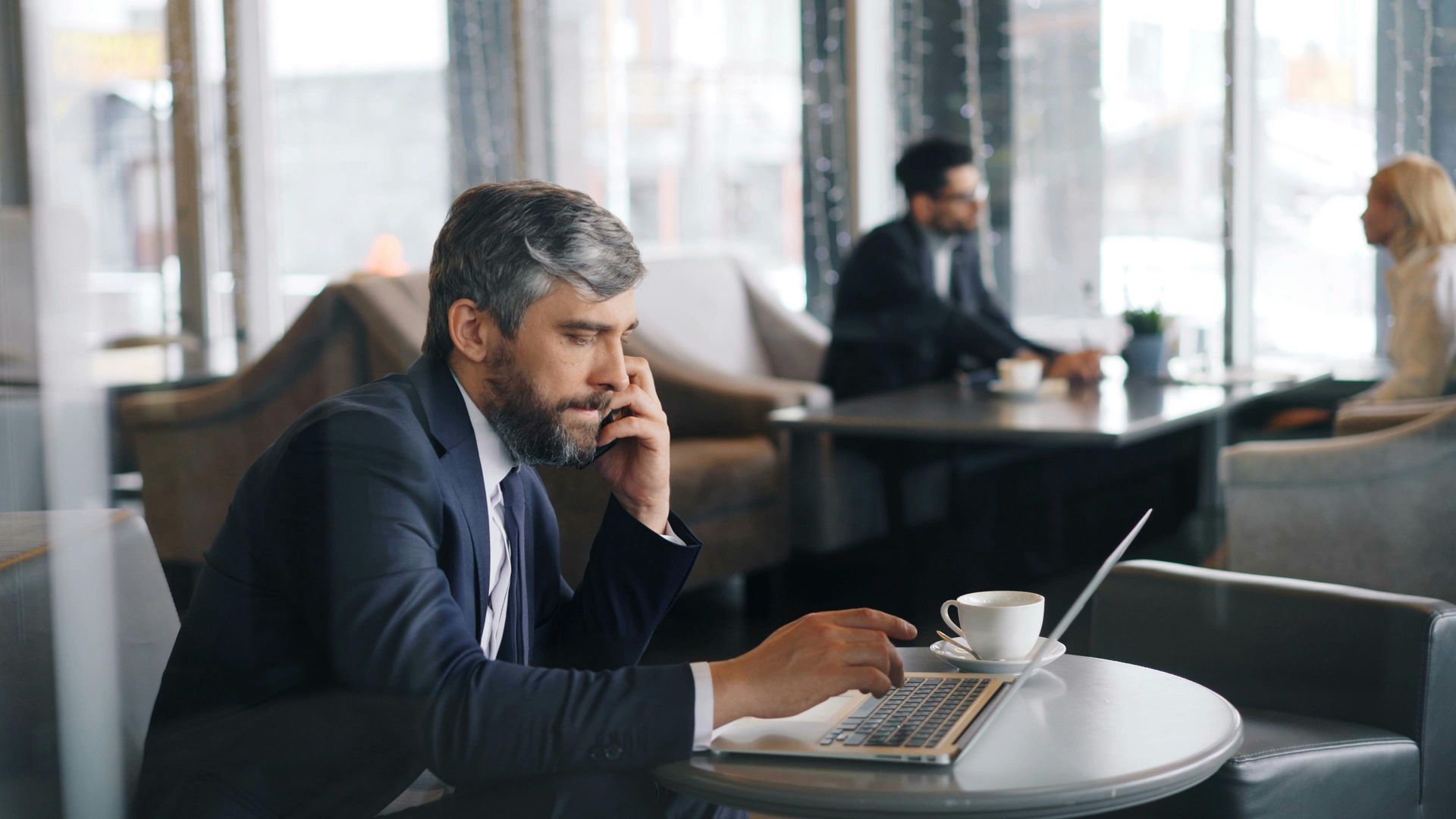 A man in a suit is sitting at a table with a laptop and talking on a cell phone.