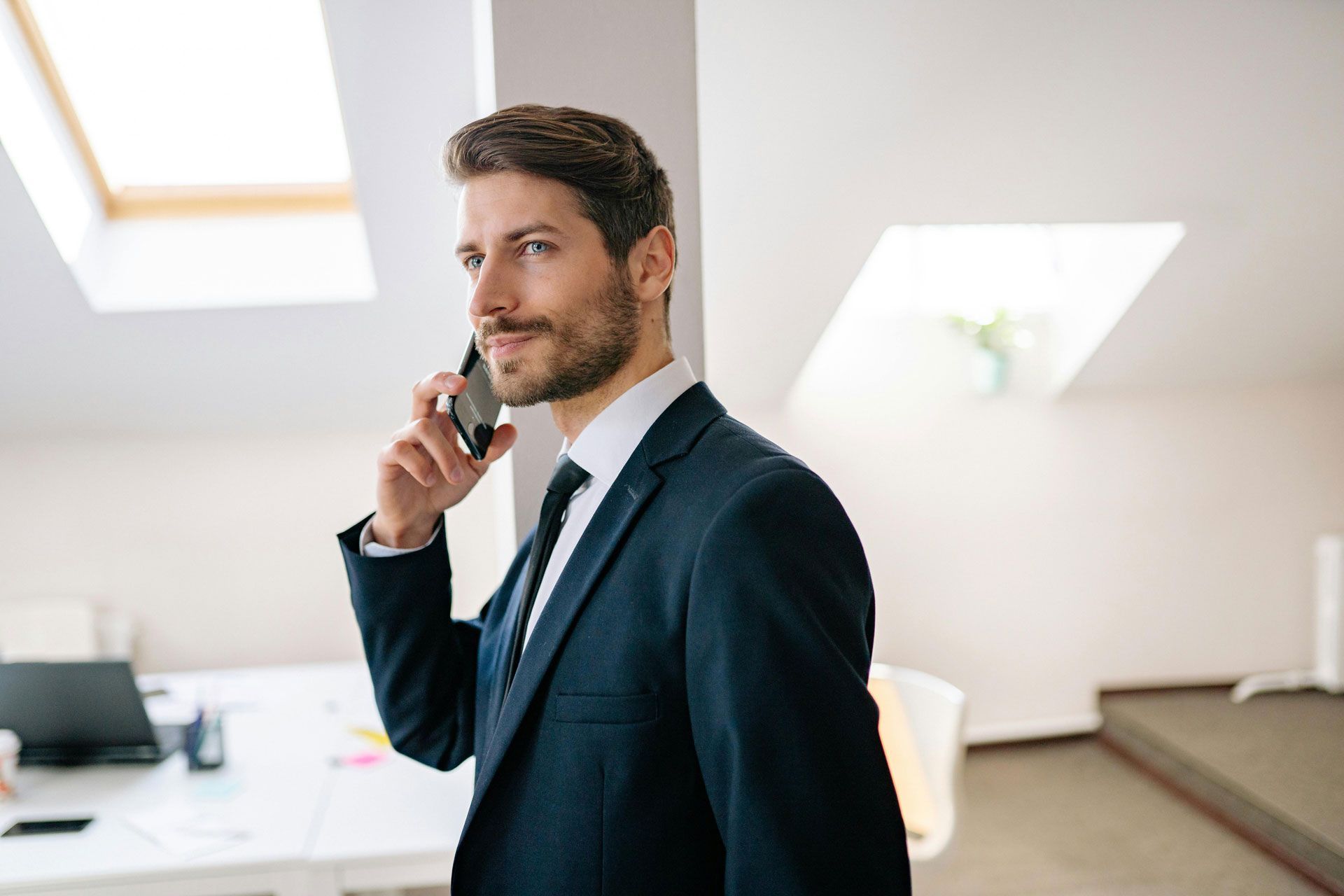 Man in a suit, talking on a phone, looking away. In a bright office, near a window.