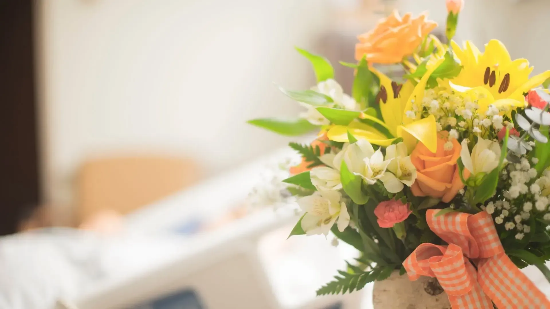 Bouquet of yellow and orange flowers with ribbon, bedside table in a soft-lit hospital room.