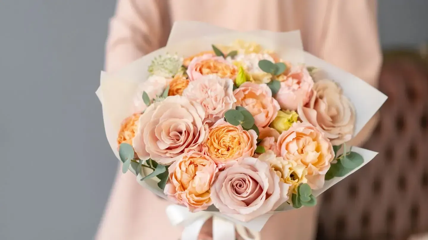 Person holding a bouquet of peach and light pink roses, wrapped in paper, tied with a ribbon.