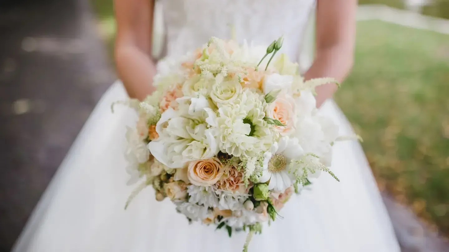 Bride in white dress holding a bouquet of cream, peach, and green flowers.